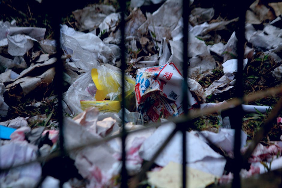 A close-up image showing a pile of discarded rubbish behind a black metal fence, consisting of crumpled paper, plastic packaging, and food wrappers scattered on the ground. The trash includes a yellow