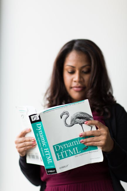 A woman with dark hair, dressed in a black blazer and maroon top, is standing indoors against a plain light-colored wall. She is focused on reading a technical book titled 'Dynamic HTML' published by